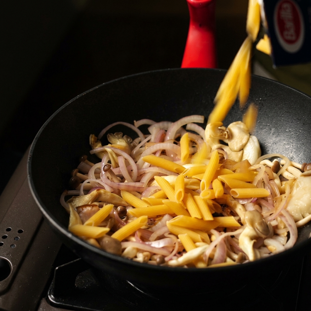 Trockene Penne-Nudeln werden aus einer Packung in eine schwarze Pfanne mit angebratenen Pilzen und Zwiebeln geschüttet.