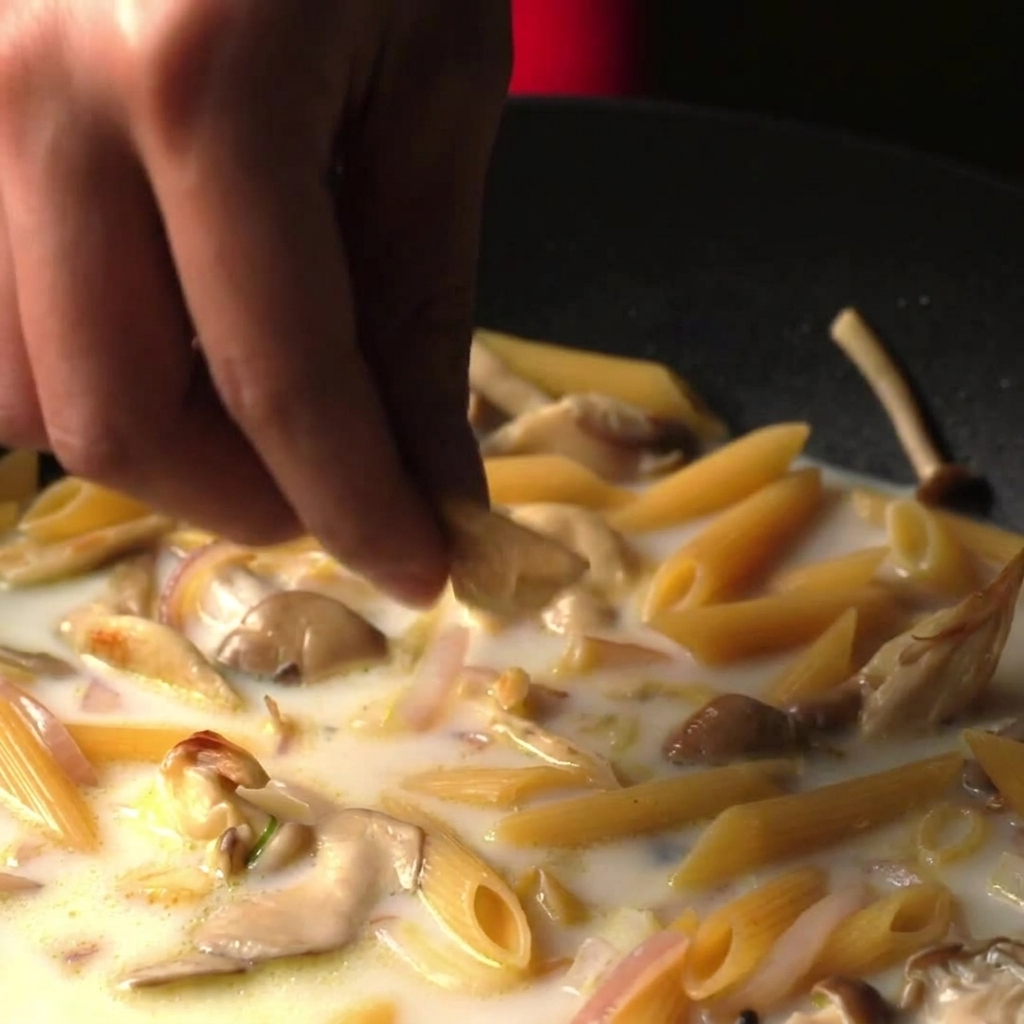 Finger lassen einen Brühwürfel in eine Pfanne mit Penne, die in einer cremigen weißen Brühe köcheln.