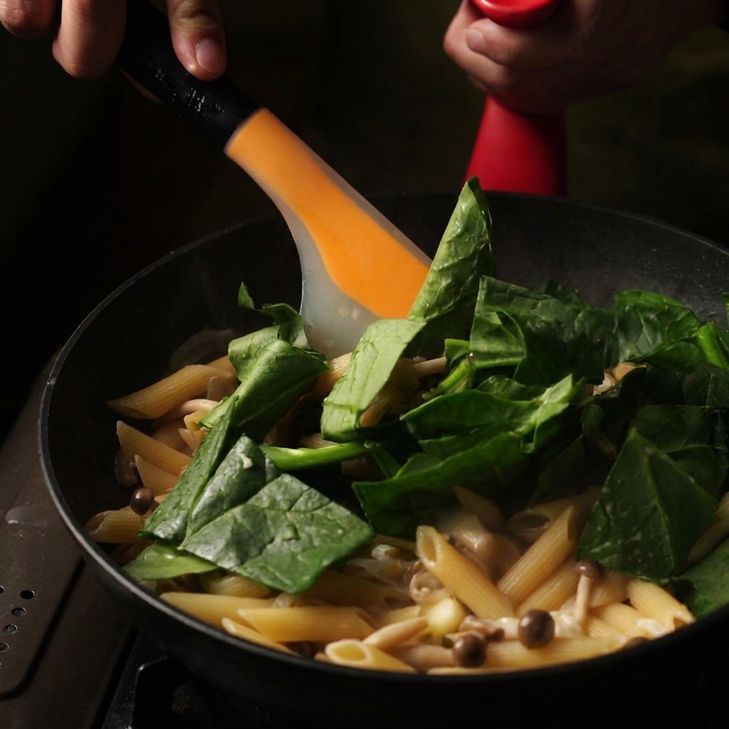 Ein orangefarbener Pfannenwender rührt frische grüne Spinatblätter und Butter in eine Pfanne mit cremiger Pasta.