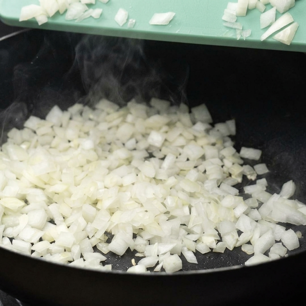 Gewürfelte weiße Zwiebeln werden von einem grünen Schneidebrett in eine dampfende schwarze Pfanne geschabt.