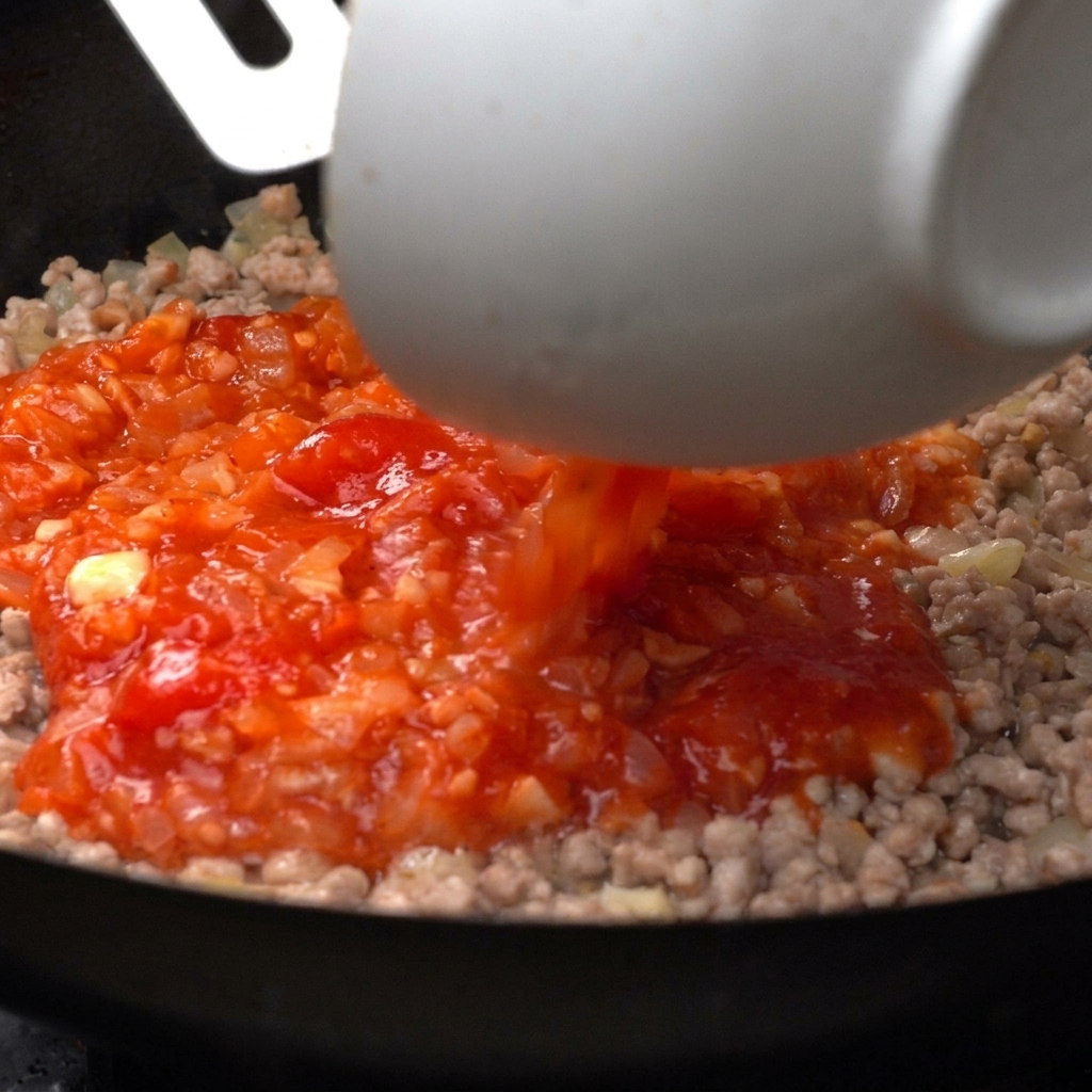 Leuchtende gestückelte Tomaten werden aus einer weißen Schüssel in eine Pfanne mit gebratenem Hackfleisch gegossen.
