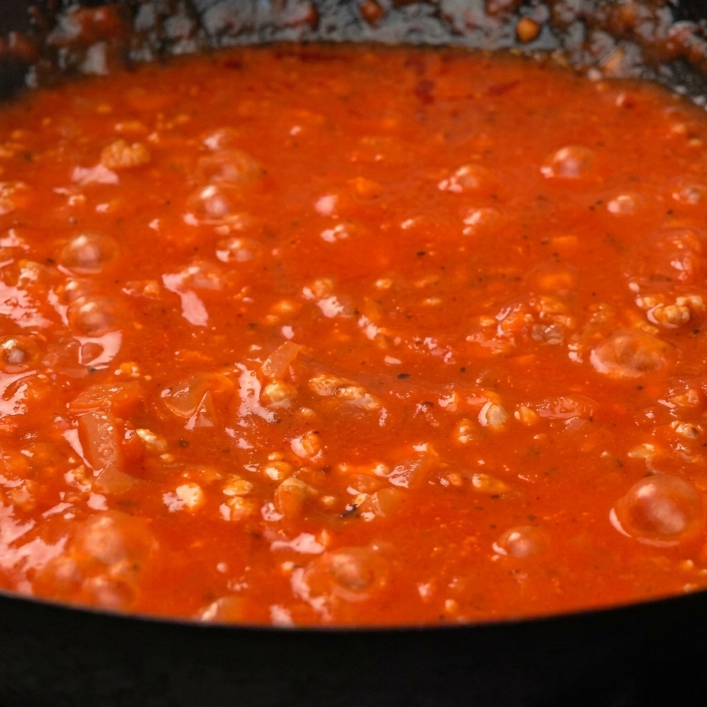 Gekochte Paccheri-Pasta wird in eine dicke, leuchtend rote Tomaten-Hackfleischsauce in einer Pfanne gerührt.