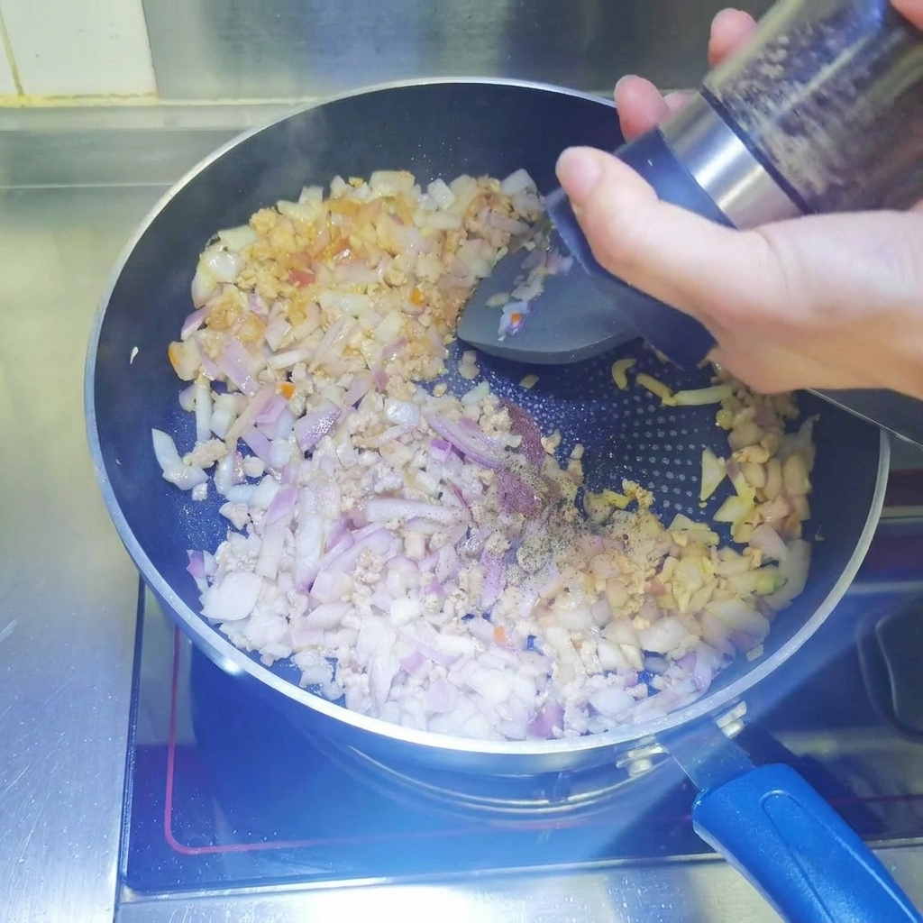 Schwarzer Pfeffer wird frisch aus einer Mühle in eine Pfanne mit bräunendem Hackfleisch und gewürfelten Zwiebeln gemahlen.