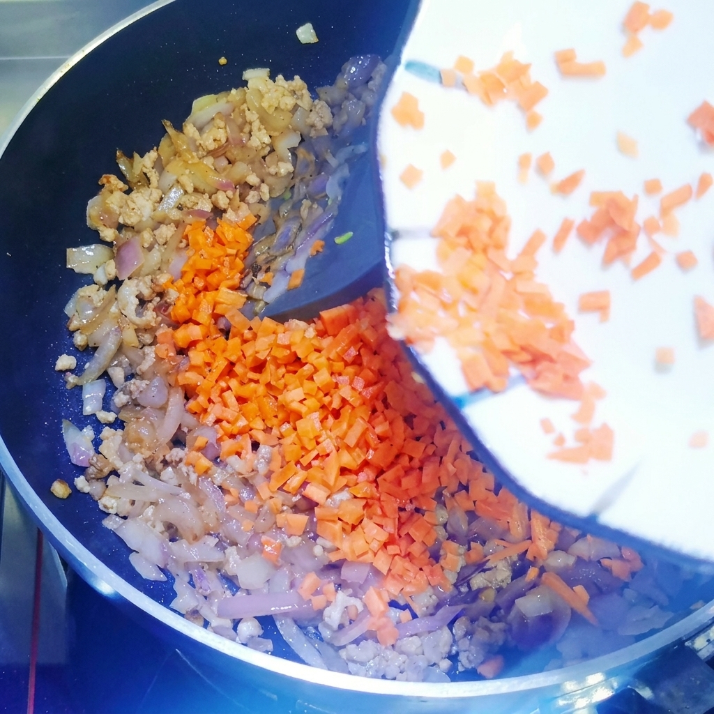 Fein gewürfelte Karotten werden in eine Pfanne mit bratendem Fleisch und Zwiebeln gegeben und mit einem Silikonspatel vermengt.
