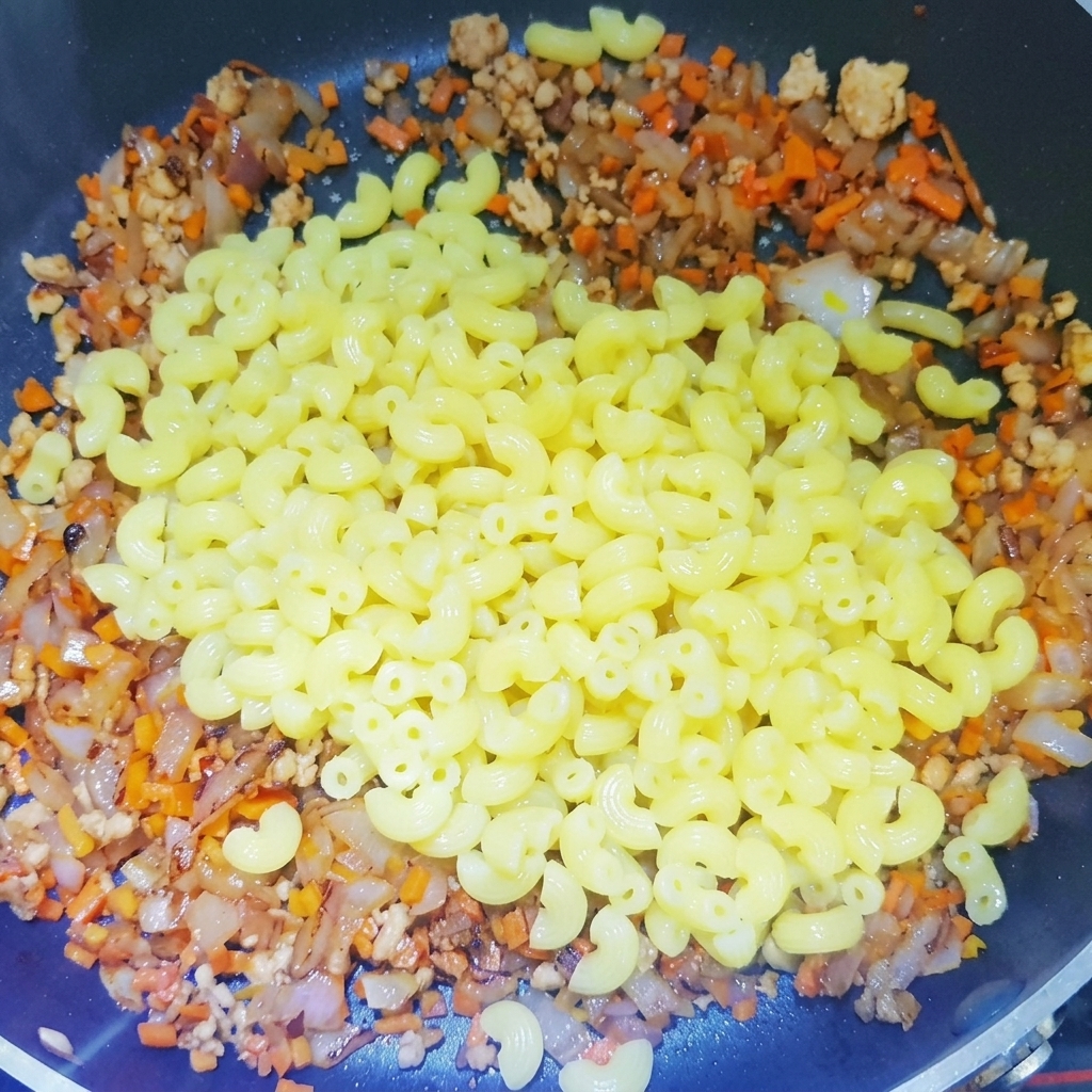 Gekochte Makkaroni werden in eine Pfanne gegeben, die mit einer fertigen Basis aus Hackfleisch, Zwiebeln und Karotten gefüllt ist.