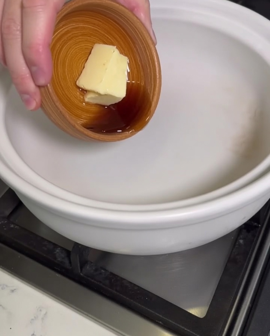Eine Hand gibt einen Block Butter und flüssiges Sesamöl aus einer kleinen Holzschale in einen leeren Tontopf.