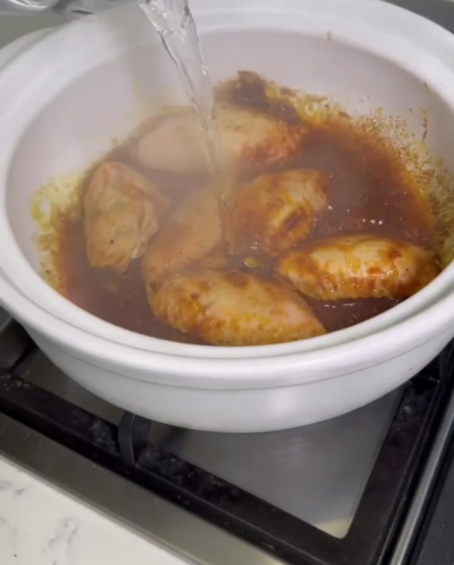 Klares Wasser wird aus einem Gefäß in einen blubbernden Tontopf voller Curry-Hähnchenflügel gegossen.