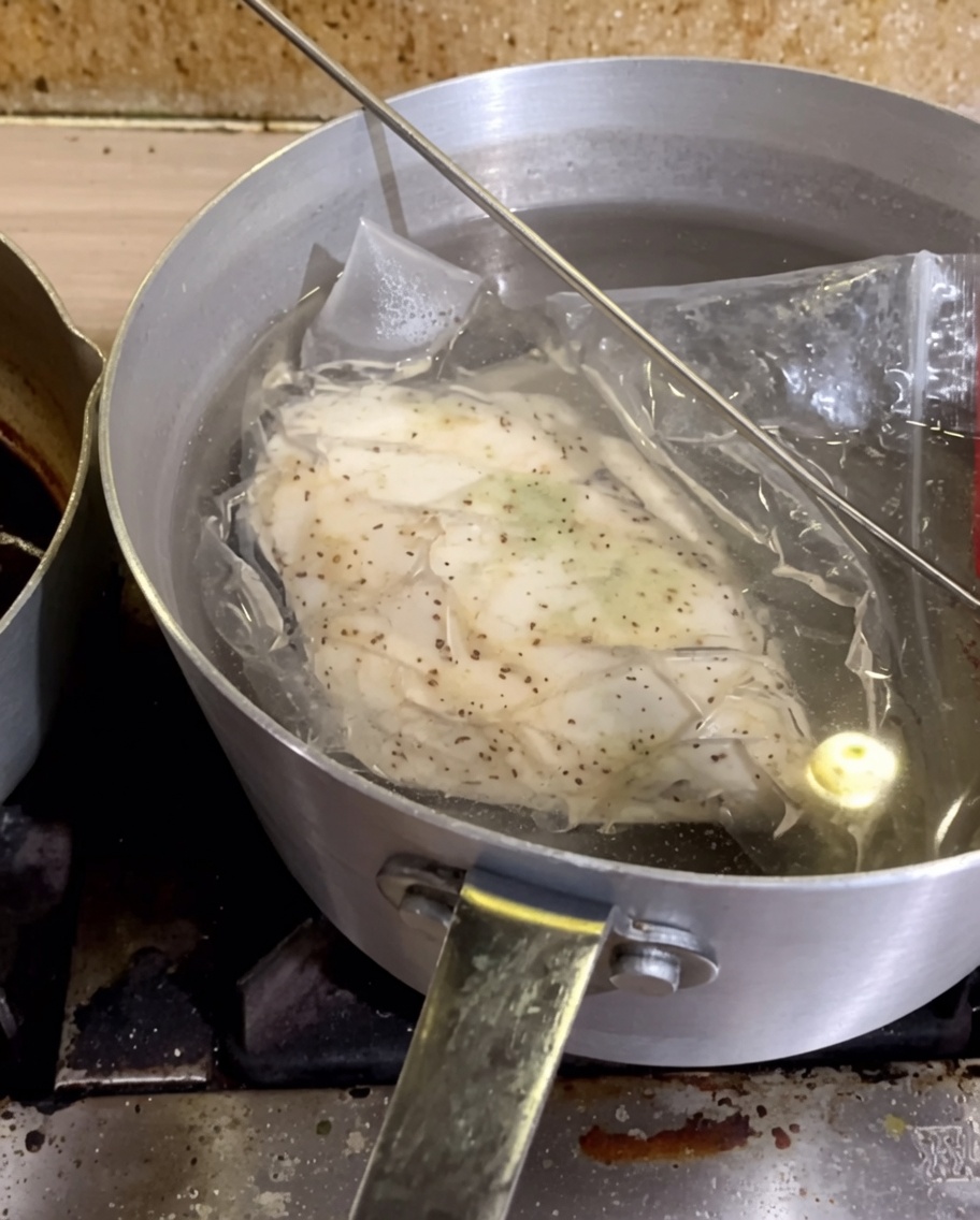 Ein vakuumierter Beutel mit gewürzter Hähnchenbrust, der in einem Topf mit heißem Wasser liegt.
