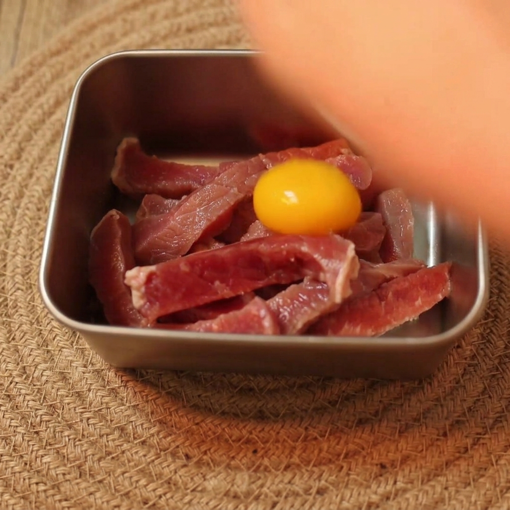 Ein hellgelbes, rohes Eigelb liegt auf in Streifen geschnittenem rohem Rindfleisch in einer Metallschüssel.