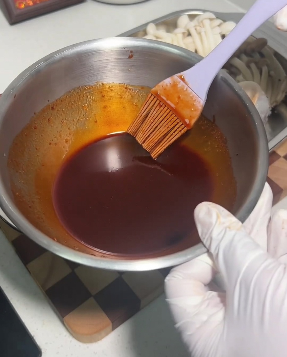 Eine Metallschüssel mit einer dunklen, würzigen Saucenmischung, die mit einem Silikonpinsel gerührt wird.