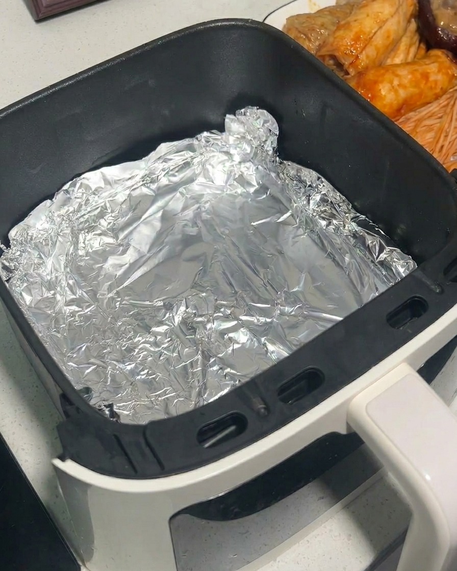 Marinierte Pilze werden mit einer Metallzange in einen mit Alufolie ausgelegten Heißluftfritteusen-Korb gelegt.