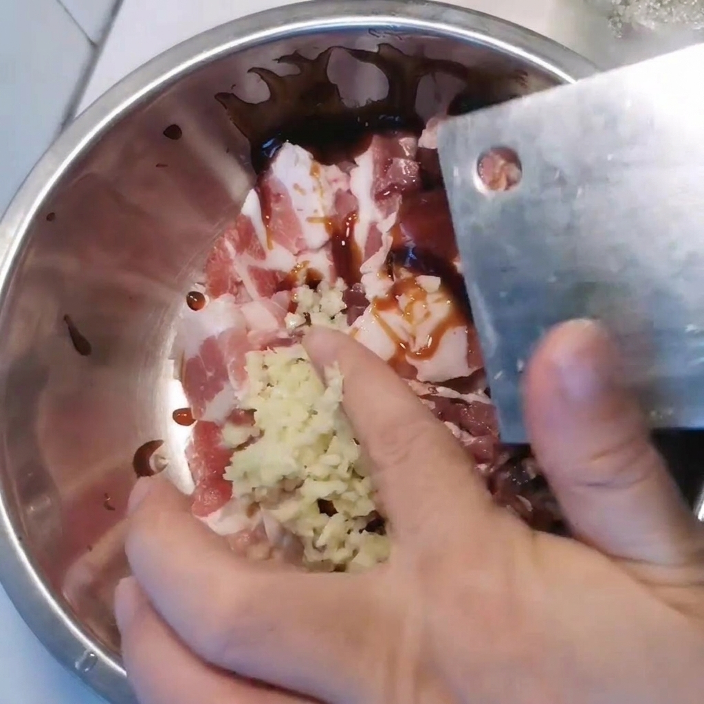 Eine Metallschüssel mit in Scheiben geschnittenem Schweinebauch, der mit gehacktem Knoblauch und verschiedenen Saucen gewürzt wird.