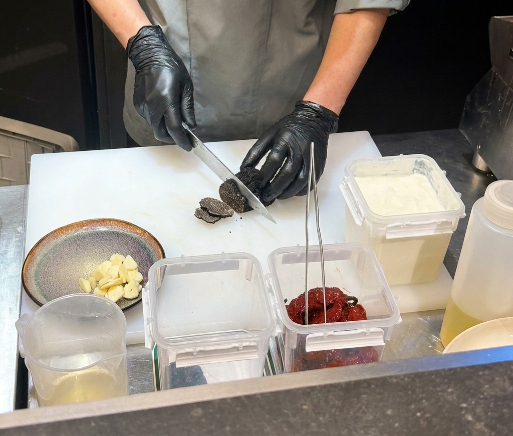 Ein Koch mit schwarzen Handschuhen hackt getrocknete schwarze Chilis auf einem weißen Schneidebrett.