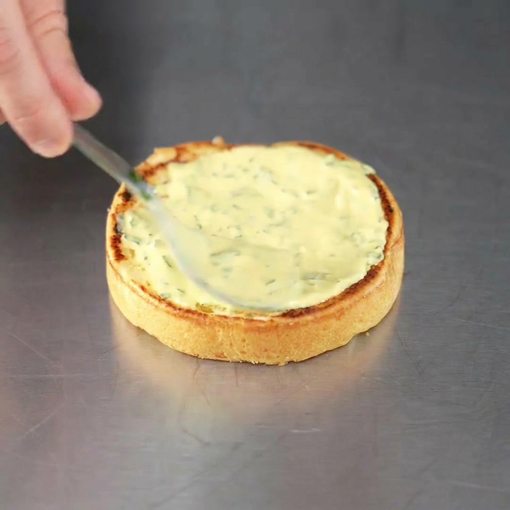 Ein frisch geröstetes Burgerbrötchen wird mit grüner Kräutersauce bestrichen und ist bereit für das Patty.