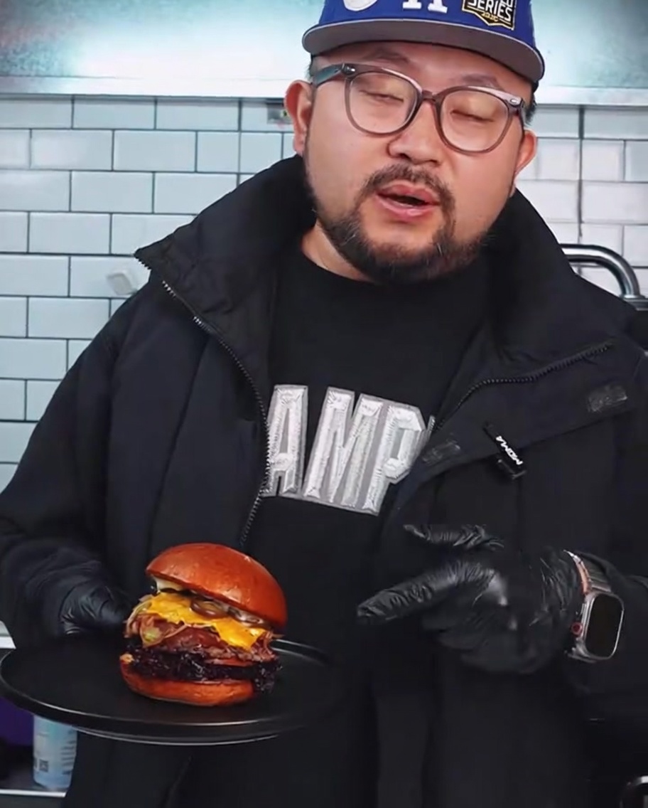 Ein rohes Wagyu-Rindfleisch-Patty wird mit einer schweren Burgerpresse aus Metall flach auf einen heißen Grill gedrückt.