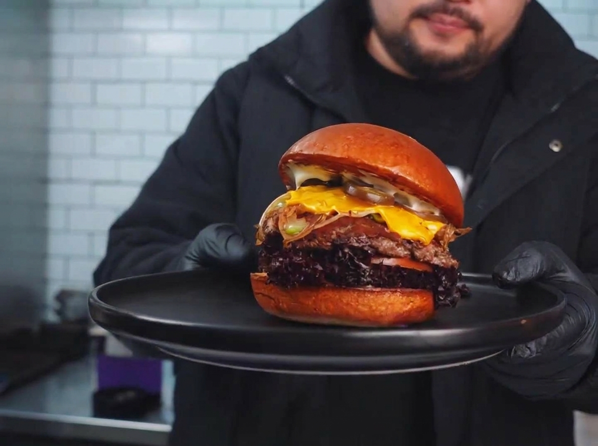 Ein gebratenes Patty auf dem Grill, belegt mit angebratenem Gemüse und einer schmelzenden Käsescheibe.