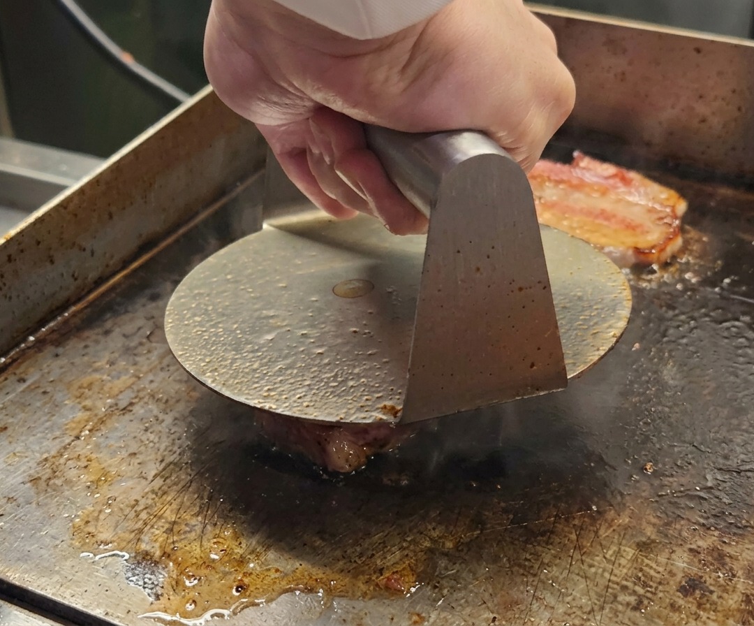 Eine Burgerpresse aus Metall flacht eine Kugel Wagyu-Hackfleisch auf einer heißen Fläche ab.