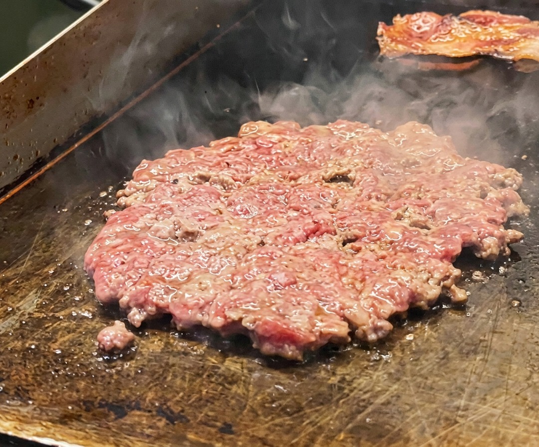 Ein dünnes Rindfleisch-Patty auf dem Grill, gewürzt mit Salz und belegt mit Zwiebelscheiben.