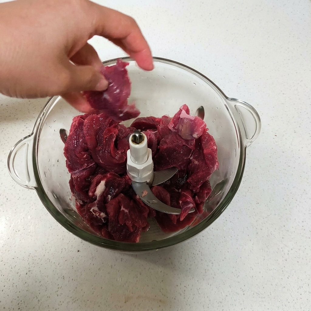 Eine Hand gibt Stücke von rohem, rotem Rindfleisch in eine Glasschüssel einer Küchenmaschine.