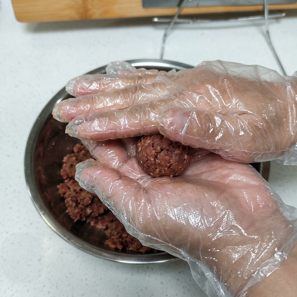 Behandschuhte Hände halten und pressen eine Kugel aus gewürztem Hackfleisch in eine Patty-Form.