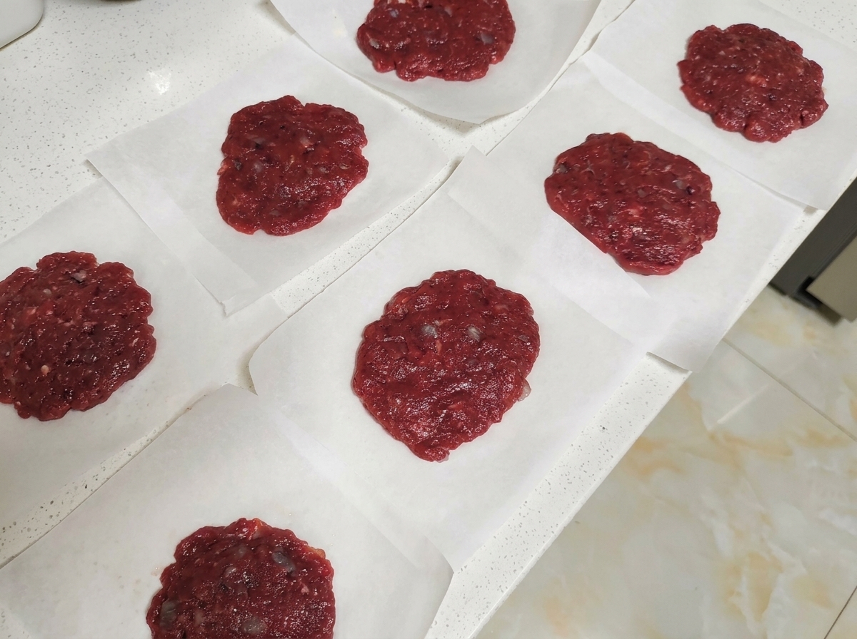 Mehrere rohe Rindfleisch-Patties liegen einzeln auf quadratischen Stücken Backpapier auf einer Arbeitsplatte.
