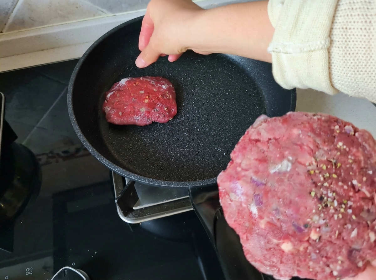 Eine Hand hält ein rohes Rindfleisch-Patty, während ein anderes in eine heiße, leicht geölte beschichtete Pfanne gelegt wird.