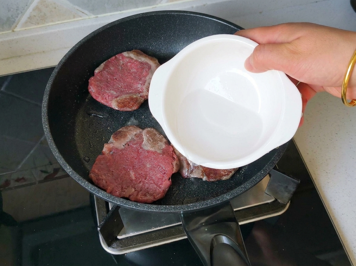 Eine Hand gießt einen Spritzer Wasser aus einer kleinen weißen Schüssel in eine Pfanne mit drei gebräunten Rindfleisch-Patties.