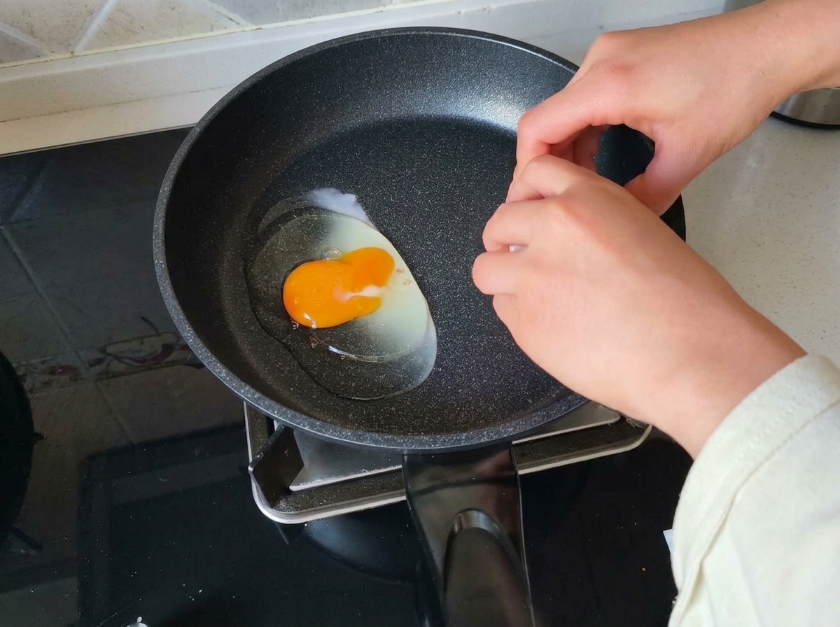 Eine Hand schlägt ein rohes Ei mit einem leuchtend gelben Dotter in eine dunkle, erhitzte Pfanne.