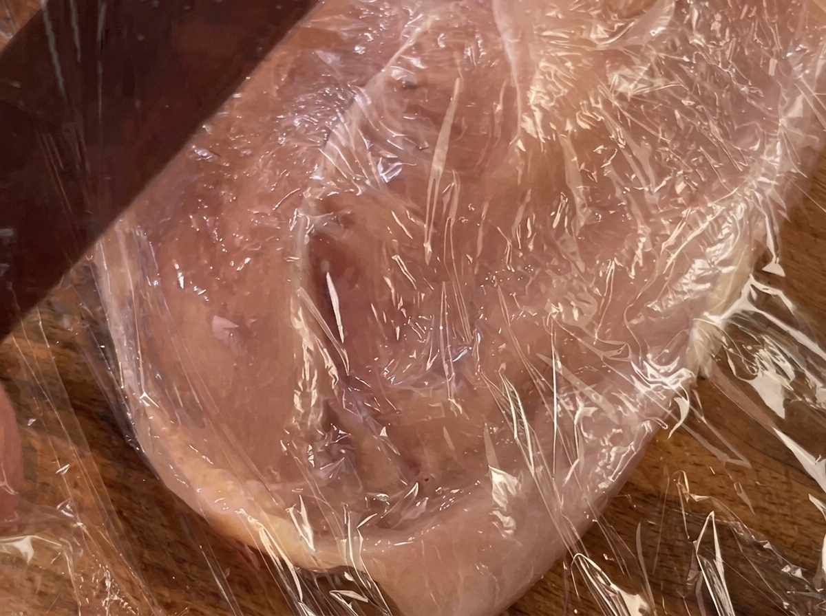 Ein Nudelholz drückt auf ein mit Plastikfolie abgedecktes Hähnchenbrustfilet.