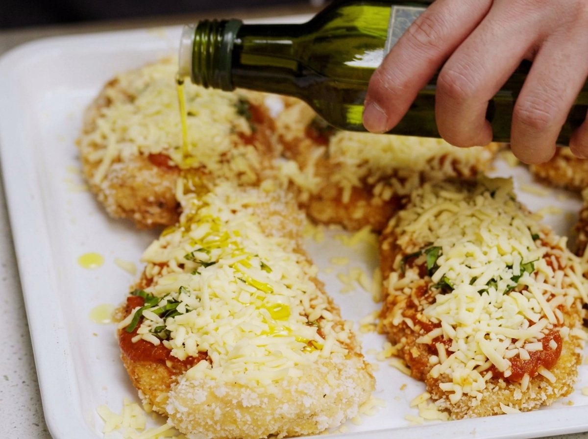 Olivenöl wird über das fertige Chicken Parmesan geträufelt.