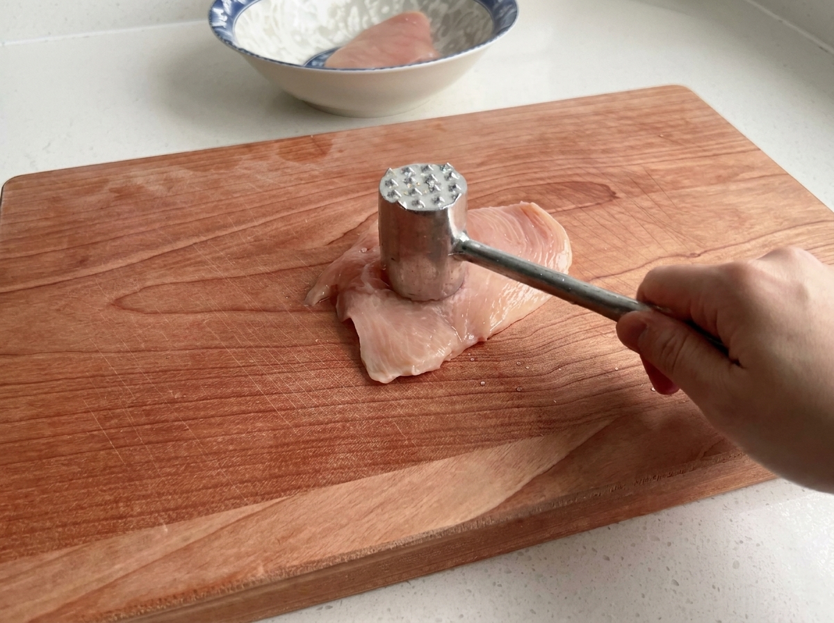 Ein Fleischklopfer bearbeitet eine rohe Hähnchenbrust auf einem Holzbrett.