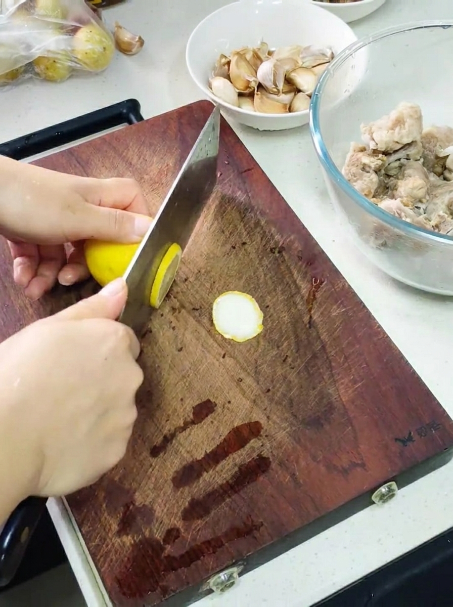 Hände schneiden mit einem Hackmesser eine Zitrone auf einem Holzbrett, daneben eine Schüssel mit marinierten Rippchen und Knoblauchzehen.