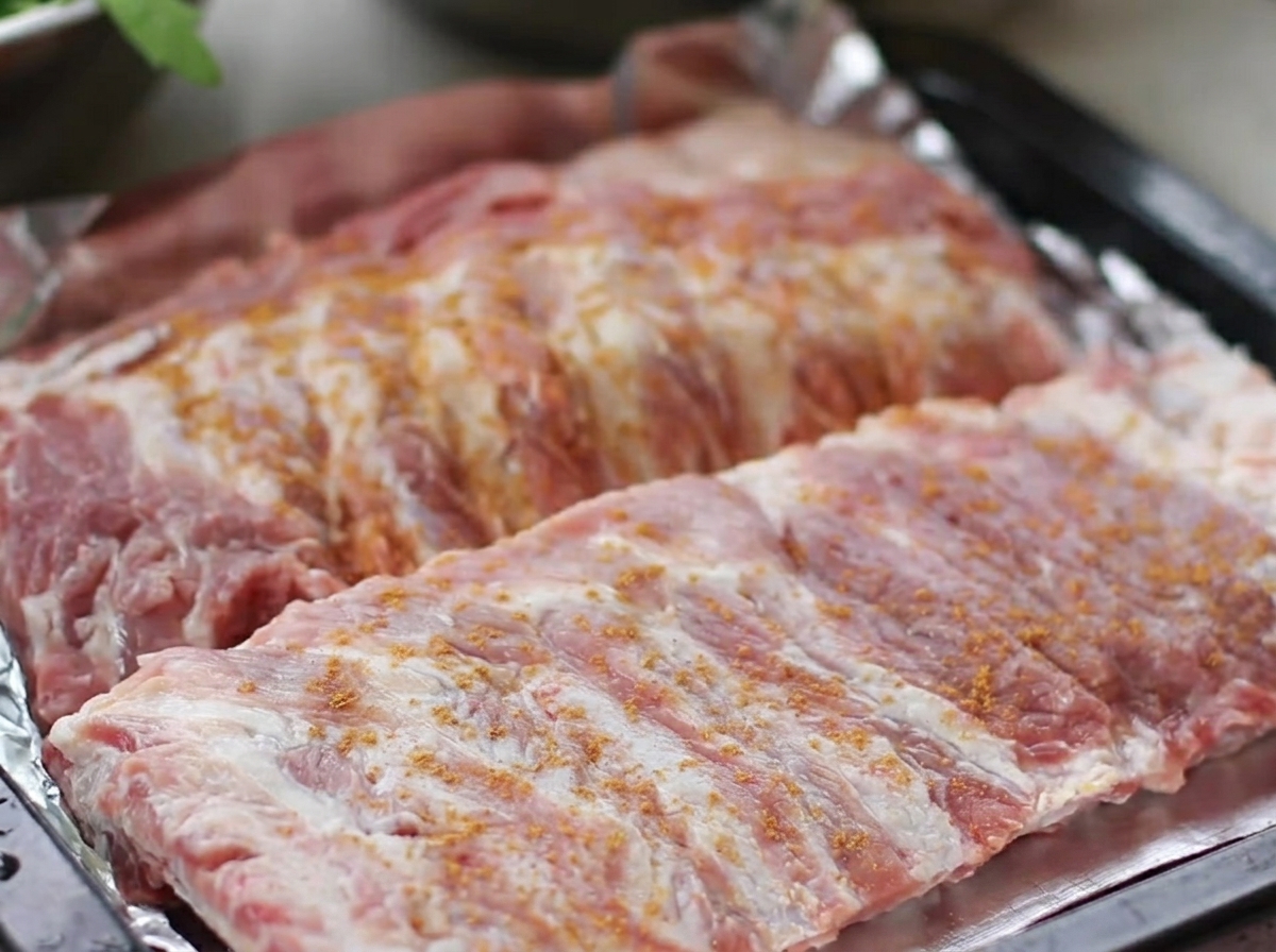 Rohe Schweinerippchen auf einem Backblech, großzügig bestreut mit einem Rub aus Fünf-Gewürzen, schwarzem Pfeffer, Ingwer und Knoblauch.