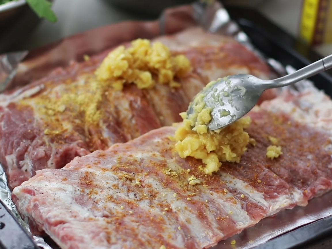 Hände massieren eine Mischung aus gehacktem Knoblauch und Gewürzen in die Oberfläche roher Rippchen auf einem Blech.