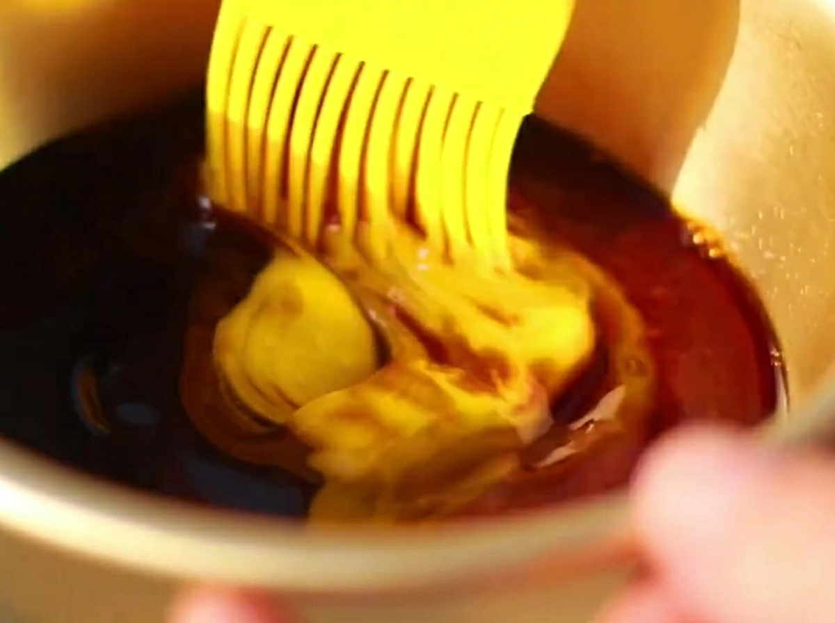 Ein Silikonpinsel trägt eine dunkle, reichhaltige Sauce auf die Oberfläche der bereits vorgegarten Rippchen auf.