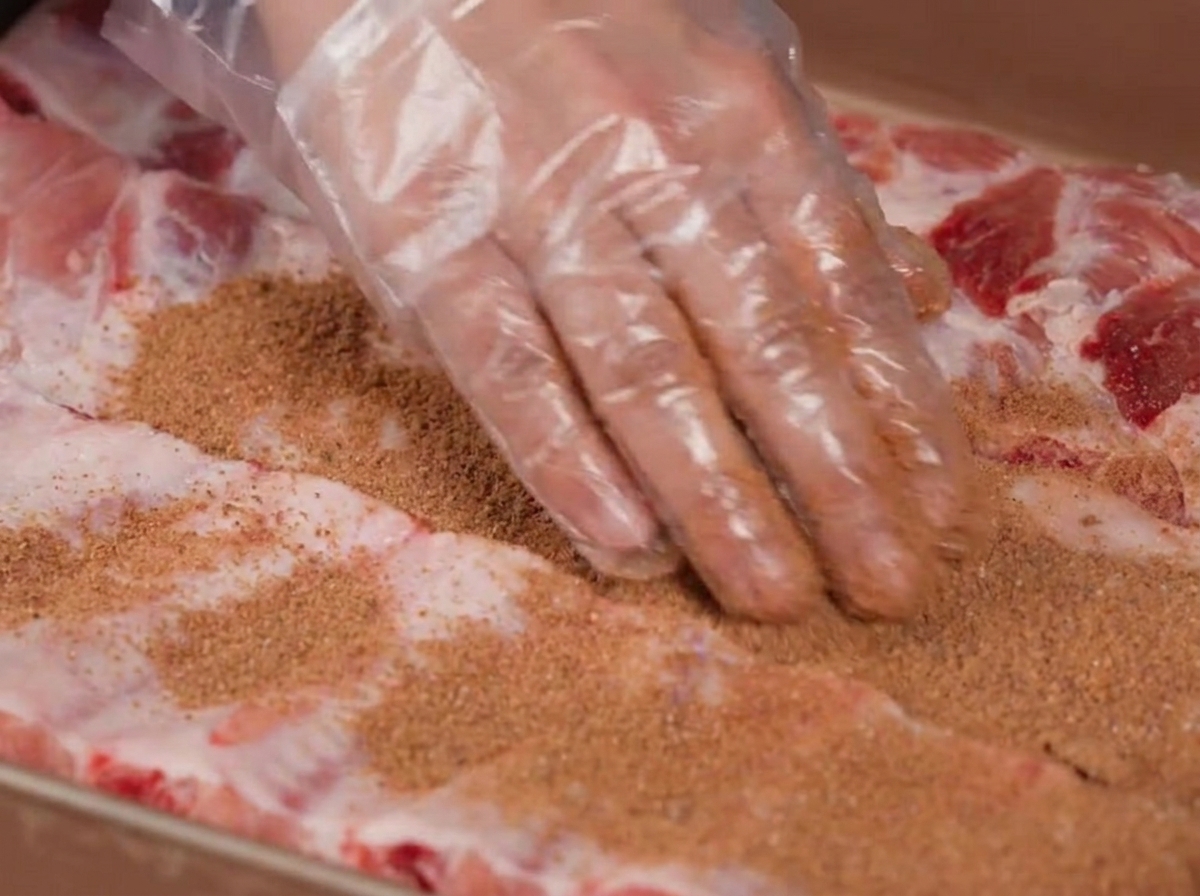 Hände in Plastikhandschuhen reiben eine braune Gewürzmischung über rohe Schweinerippchen.