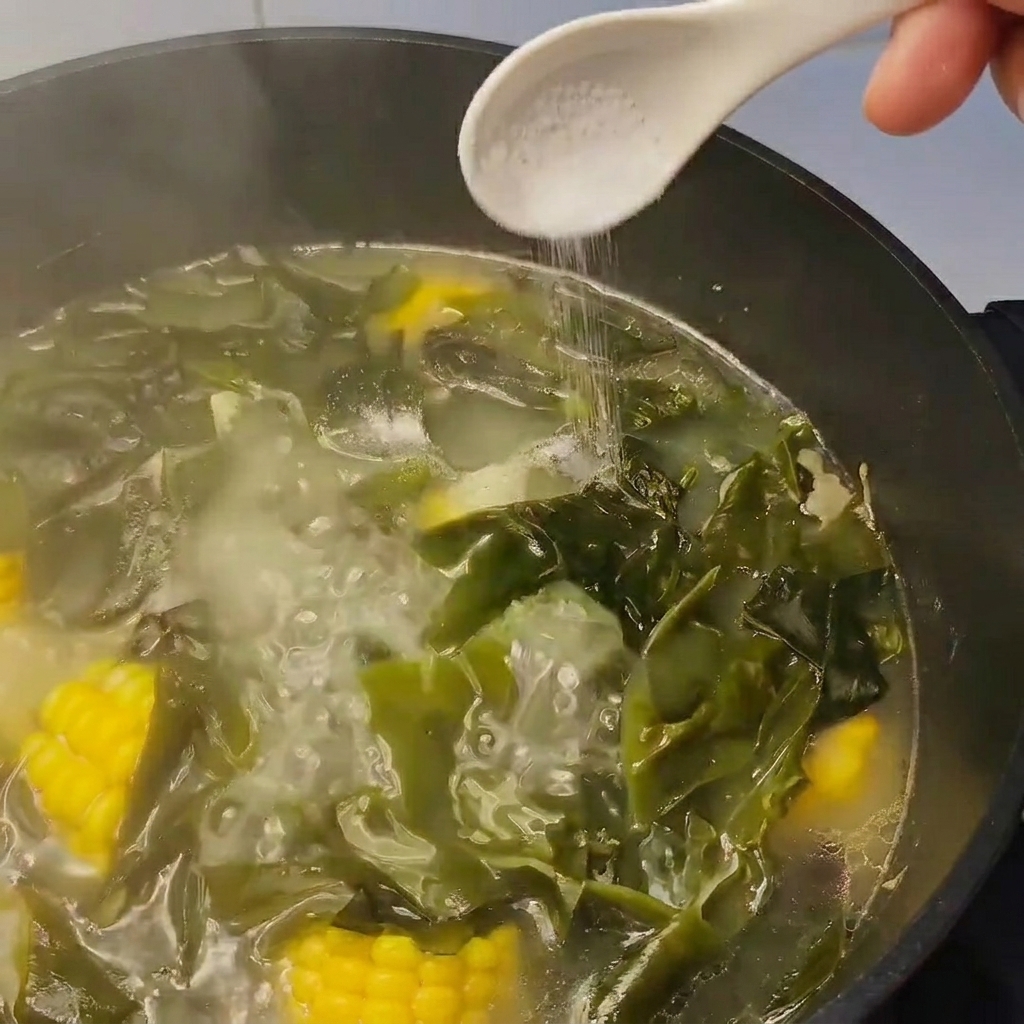 Ein Löffel Salz wird in einen köchelnden Suppentopf mit Kelp und Mais gegeben.