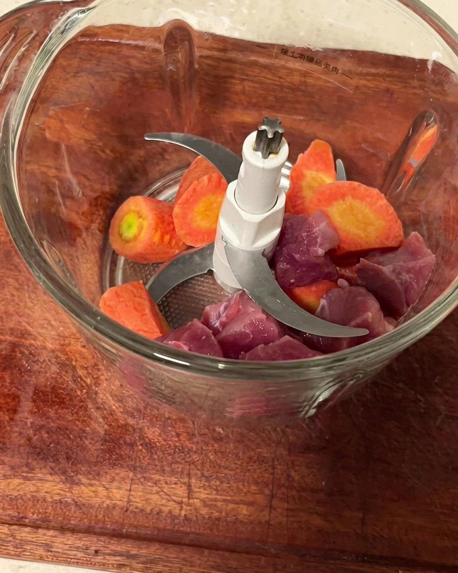 Schweinefleischstücke und Karottenwürfel in einem Fleischwolfbehälter bereit zum Zerkleinern.