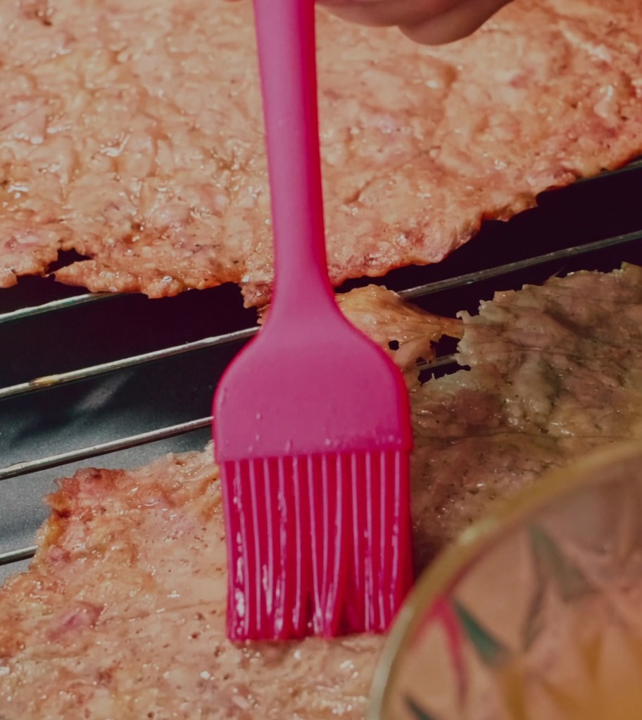 Eine Silikonpinsel verteilt eine Honig-Wasser-Glasur auf Schweinefleischscheiben auf einem Drahtgitter.