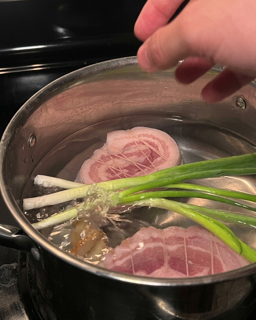 Gebundener Schweinebauch in einem Topf mit kochendem Wasser und Frühlingszwiebeln.