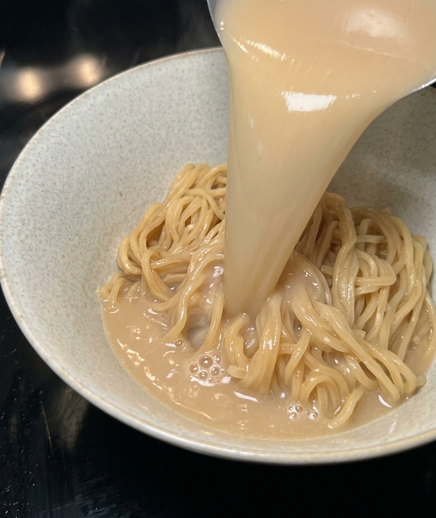 Heißer, cremiger Tonkotsu-Brühe wird über Nudeln in eine Schüssel gegossen.