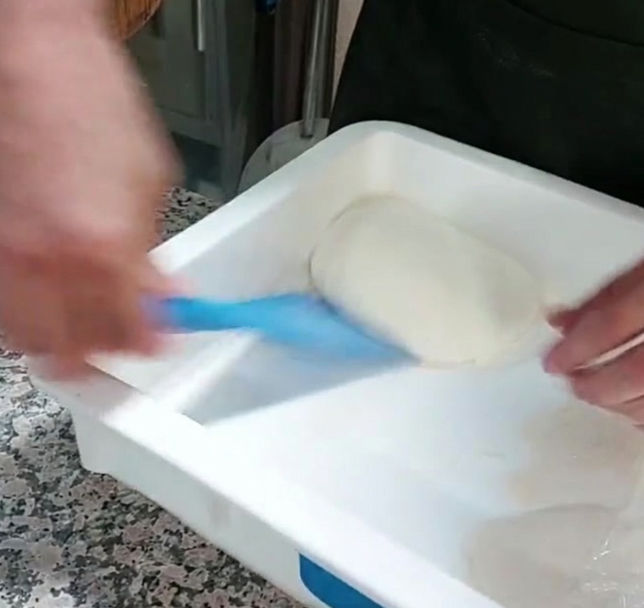 Ein Teil Pizzateig wird mit einem blauen Kunststoffschaber aus einem Behälter gehoben.