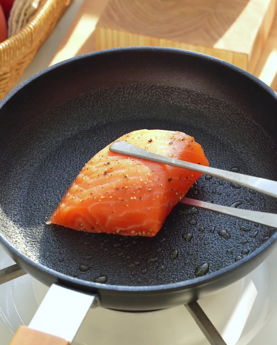 Ein gewürztes Lachsfilet wird mit einer Metallzange in eine heiße, geölte Pfanne gelegt.
