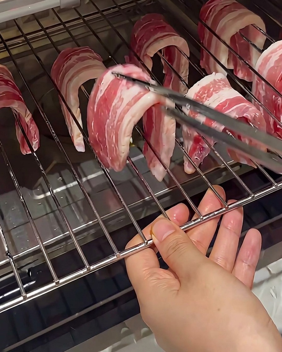Rohe Schweinebauchscheiben, die mit einer Zange über die Metallstreben eines Ofenrosts gehängt werden.