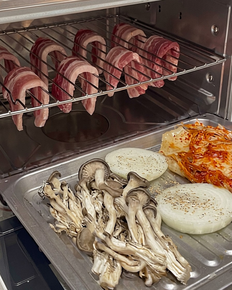 Backblech mit Gemüse und Kimchi, das im Ofen unter dem hängenden Schweinebauch positioniert ist.