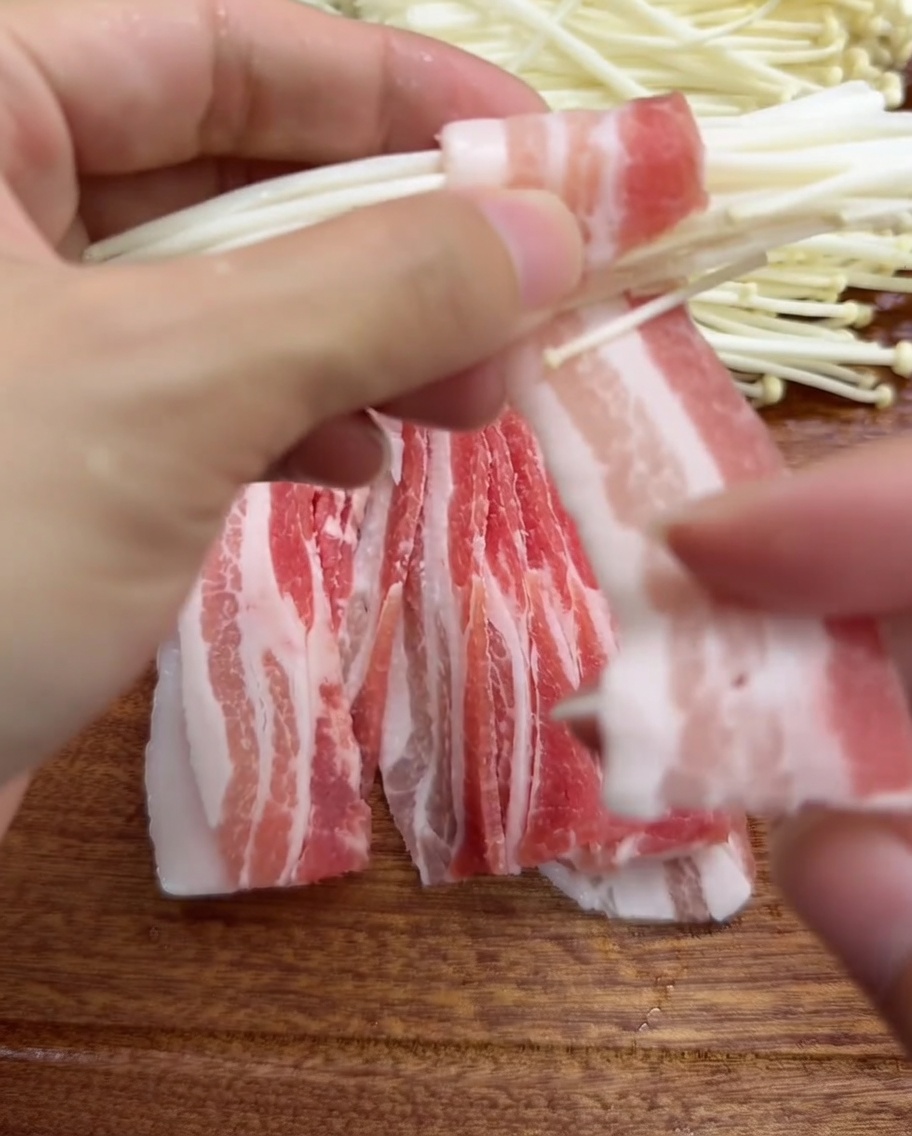 Nahaufnahme von Händen, die eine dünne Scheibe Schweinebauch um ein Bündel Enoki-Pilze wickeln.