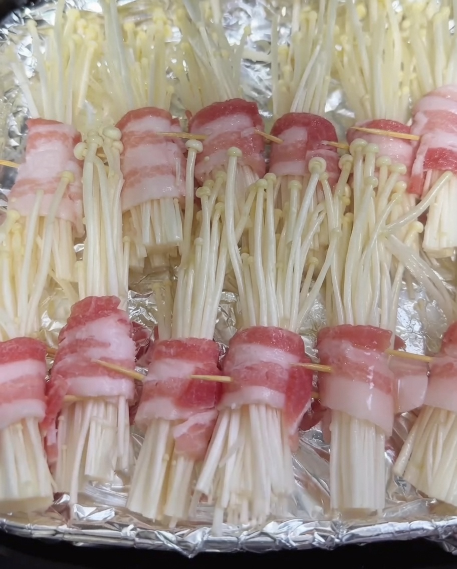 Sprühflasche beim Auftragen eines feinen Ölnebels auf die rohen, in Schweinebauch eingewickelten Enoki-Pilze im Air-Fryer-Korb.
