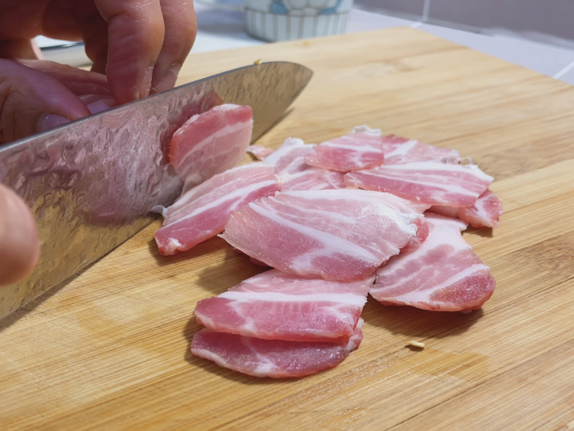 Dünne Scheiben rohen Schweinebauchs liegen auf einem Holzschneidebrett neben einem großen Kochmesser.