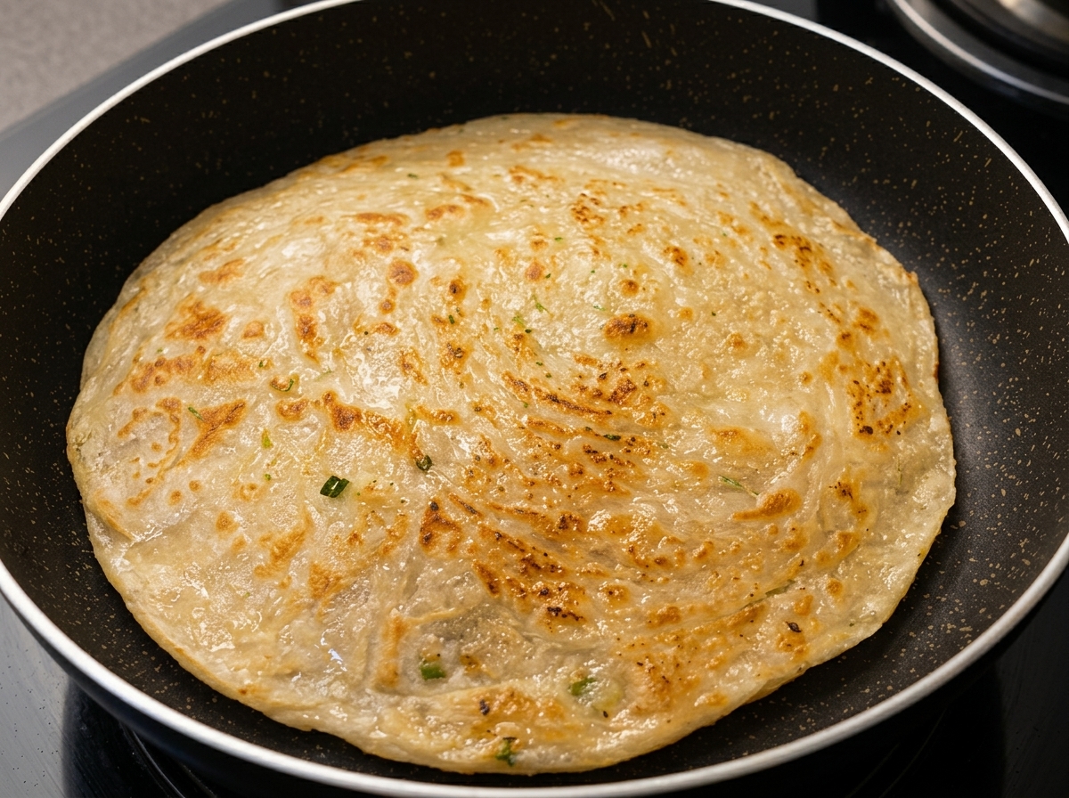 Ein rundes Frühlingszwiebel-Fladenbrot brät in einer dunklen Antihaftpfanne und präsentiert eine goldbraune, strukturierte Oberfläche.