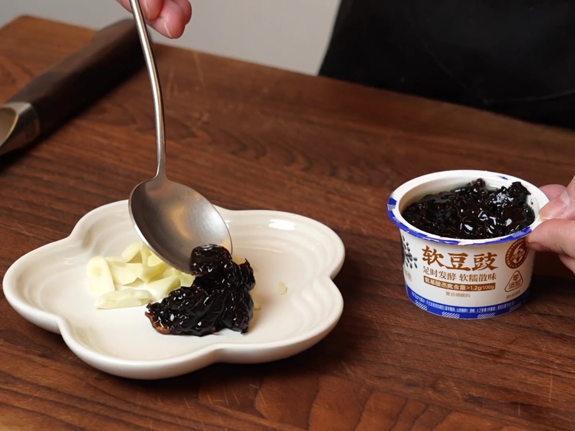 Ein Löffel schöpft feuchte fermentierte schwarze Bohnen aus einem Behälter auf einen kleinen weißen Teller, auf dem bereits Knoblauch liegt.